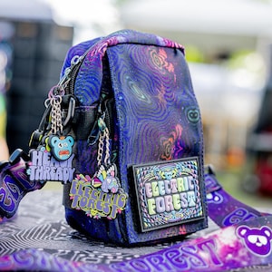 May include: A purple and blue crossbody bag featuring a cosmic pattern, a rectangular Electric Forest patch, and multiple colorful character charms attached to the zipper. The adjustable strap displays the text Electric Forest with bear icons.