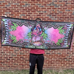 May include: A rectangular black beach towel with a vibrant flamingo and tropical design. The towel features the text "OKEECHOBEE" and "Heady Threads" in a decorative font, with a pink, purple, and blue color scheme. Palm trees and floral elements surround the central image.
