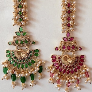 Może przedstawiać: Dwa złote, bogato zdobione kolczyki. Jeden ma zielone kamienie szlachetne i perły, drugi czerwone kamienie szlachetne i perły. Każdy ma dekoracyjny wzór pawia i zwisający łańcuszek z pereł i kamieni szlachetnych.