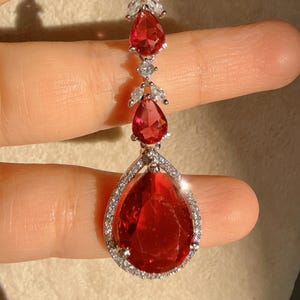 May include: A silver-toned pendant necklace featuring a series of red, teardrop-shaped gemstones. The largest gemstone is at the bottom, surrounded by a halo of small, clear stones. The necklace is held up by a hand.