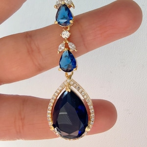 Może przedstawiać: Złoty naszyjnik z wisiorem w kształcie łzy, który zawiera duży niebieski szafir otoczony mniejszymi, przezroczystymi kamieniami. Wisiorek jest zawieszony na łańcuszku z mniejszych niebieskich szafirów i przezroczystych kamieni.