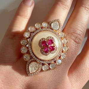 May include: A large, ornate ring with a gold band and a central cream-colored stone. The ring features a cluster of small, red gemstones in the center, surrounded by clear, round and pear-shaped stones. The ring is set with many small, clear gemstones.