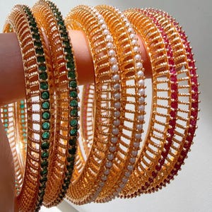 May include: Set of five gold bangles with decorative designs. Two bangles feature green gemstones, one has white pearls, and two have red gemstones.