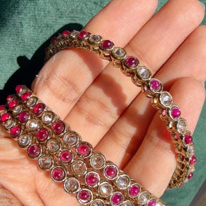 May include: Gold-toned bangles adorned with alternating red and clear gemstones. The jewelry features a repeating pattern of circular stones set in a gold metal band. The bangles are displayed against a green background.