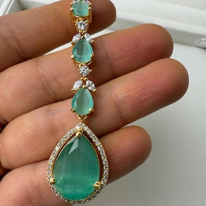 Może przedstawiać: Złoty naszyjnik z wisiorem w kształcie łzy z zielonego kamienia szlachetnego otoczonego mniejszymi, przezroczystymi kamieniami szlachetnymi. Naszyjnik składa się z szeregu mniejszych, zielonych kamieni szlachetnych w kształcie łzy z przezroczystymi kamieniami szlachetnymi po obu stronach.