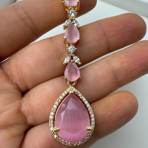 Może przedstawiać: Złoty naszyjnik z wisiorem w kształcie gruszki z różowego kamienia szlachetnego otoczonego mniejszymi, przezroczystymi kamieniami szlachetnymi. Wisiorek jest największym z pięciu różowych kamieni szlachetnych w kształcie gruszki na naszyjniku.