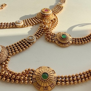 May include: Ornate gold-toned jewelry set featuring a necklace, arm cuffs, and a ring. The pieces are adorned with small beads, intricate patterns, and gemstones in red, green, and white. The set is displayed on a light-colored surface.