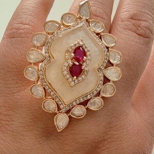 May include: A gold ring with a white stone center and two red stones. The ring is surrounded by smaller white stones.