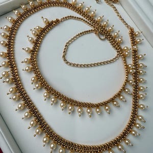 May include: A gold-coloured body chain adorned with faux pearls. The chain has a detailed design with small, hanging pearls. The chain is presented on a white surface, highlighting its intricate details and elegant style. The chain is designed to be worn around the waist.