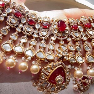 Puede incluir: Un conjunto de collar y pendientes dorados con piedras preciosas claras y rojas, y detalles de perlas colgantes. El collar tiene m&uacute;ltiples hilos con detalles intrincados y un colgante central. Las joyas se exhiben sobre un fondo claro.