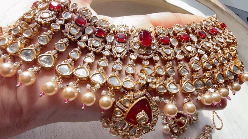 Puede incluir: Un conjunto de collar y pendientes dorados con piedras preciosas claras y rojas, y detalles de perlas colgantes. El collar tiene m&uacute;ltiples hilos con detalles intrincados y un colgante central. Las joyas se exhiben sobre un fondo claro.