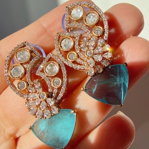 May include: A pair of gold-toned earrings with a floral design, encrusted with small diamonds and larger clear stones. Each earring features a triangular, light blue gemstone at the bottom.