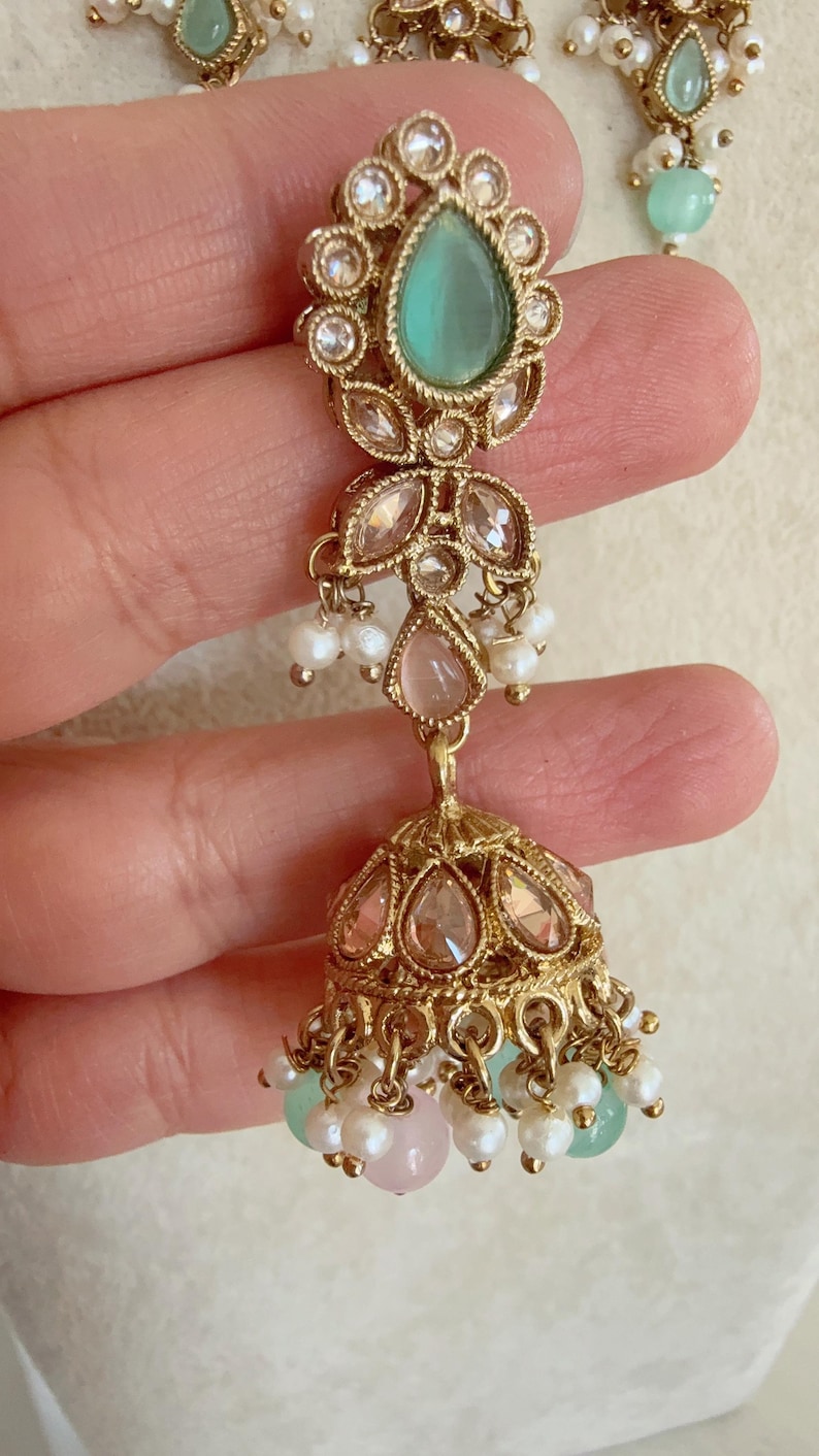 Pode incluir: Brincos dourados com uma pedra verde em forma de l&aacute;grima, p&eacute;rolas rosa e brancas e um pingente redondo dourado adornado com joias.