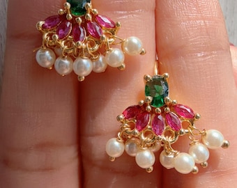 Pode incluir: Um par de brincos dourados com uma pedra verde oval central, um arranjo em leque de pedras cor-de-rosa lapidadas em marquise e uma franja pendente de pequenas pérolas brancas esféricas suspensas na base da armação metálica.