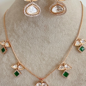 May include: A gold necklace and earrings set with a teardrop-shaped pendant and green square stones. The pendant is surrounded by smaller white stones.