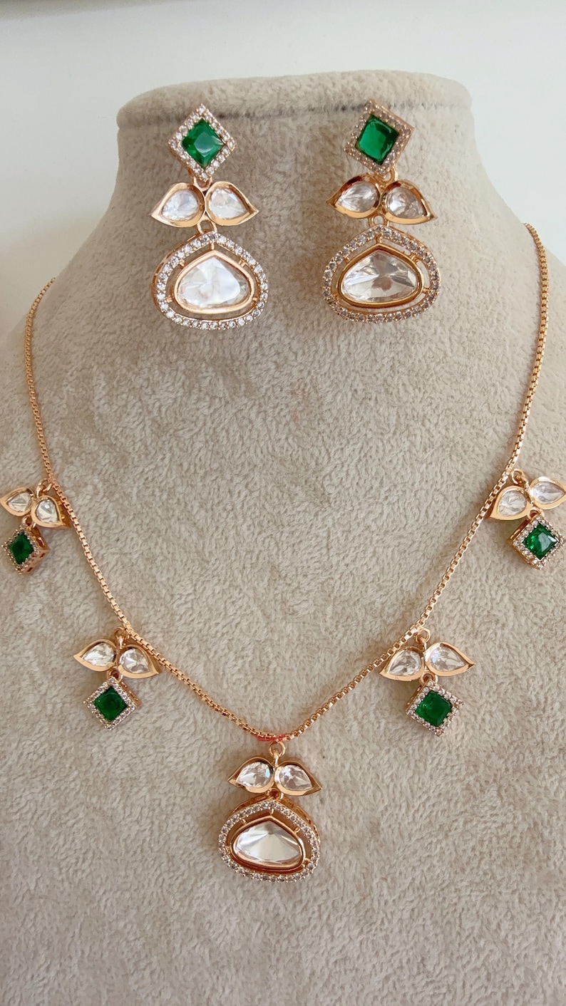May include: A gold necklace and earrings set with a teardrop-shaped pendant and green square stones. The pendant is surrounded by smaller white stones.
