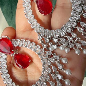 May include: A pair of silver-toned hoop earrings. Each earring features a large, teardrop-shaped, red gemstone at the top and a dangling cluster of clear, teardrop-shaped stones and small white pearls. The earrings are set against a green background.