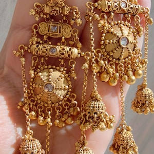 May include: Ornate gold-toned earrings with intricate detailing. Each earring features multiple levels of design, including circular and square elements, chains, and dangling bell-shaped charms. Small, clear stones are set within the design.