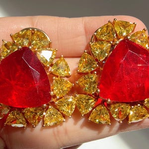 Puede incluir: Un par de pendientes de botón dorados. Cada pendiente presenta una gran gema triangular de color rojo rubí en el centro, rodeada de piedras triangulares más pequeñas de color amarillo dorado. Los pendientes están diseñados para captar la luz.