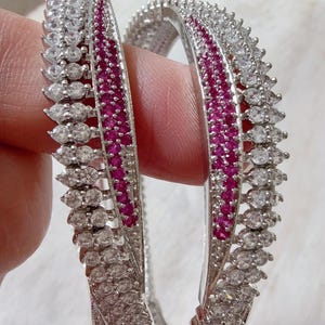 May include: Two silver bangles with a row of small, round, clear stones and a row of marquise-cut clear stones. The center of each bangle features a row of small, round, red stones. The bangles are displayed against a soft, white background.