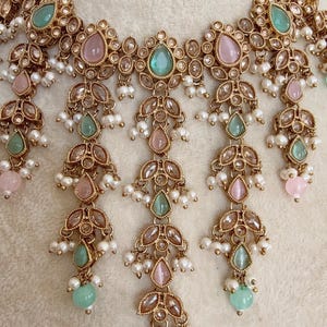 Pode incluir: Um colar de ouro com gemas rosa e verdes e detalhes de p&eacute;rolas. O colar tem um design em camadas com v&aacute;rias fileiras de p&eacute;rolas e gemas.