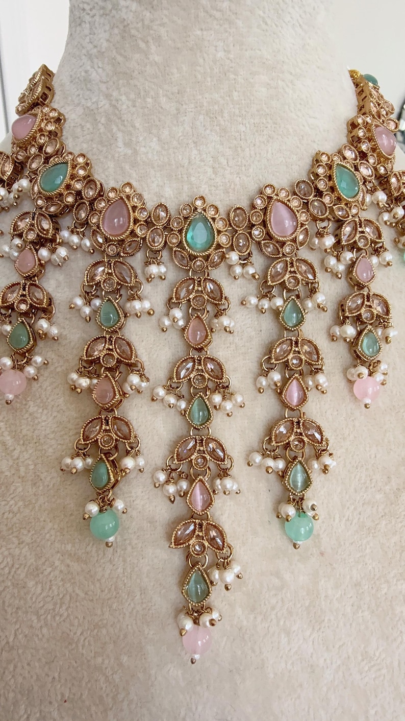 Pode incluir: Um colar de ouro com gemas rosa e verdes e detalhes de p&eacute;rolas. O colar tem um design em camadas com v&aacute;rias fileiras de p&eacute;rolas e gemas.