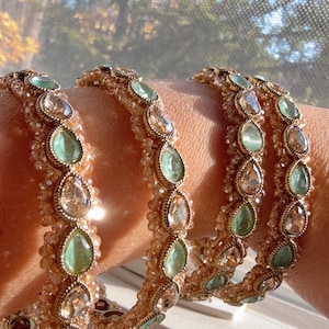 May include: Gold-toned bangles with alternating teardrop-shaped green and clear gemstones. The bracelets are adorned with small, sparkling beads, creating a luxurious and elegant look. The jewelry is perfect for adding a touch of glamour to any outfit.