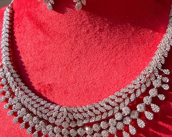 May include: A dazzling silver-toned jewelry set, including a necklace and matching earrings. The necklace and earrings feature a leaf-like design, embellished with numerous sparkling stones. The set is displayed against a vibrant red backdrop, highlighting the jewelry's brilliance.