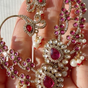 May include: Ornate gold-toned earrings with ruby-red gemstones and clear crystals. The earrings feature floral and teardrop designs, accented with small white pearls and red beads. A luxurious jewelry piece.