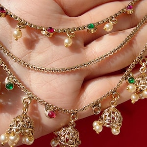 May include: Gold-toned jewelry set featuring three delicate necklaces. Each necklace is adorned with small pearls, red and green gemstones, and bell-shaped pendants. The jewelry is displayed against a soft background.
