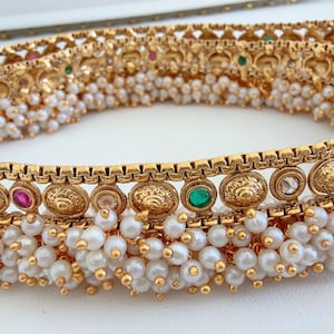 Könnte beinhalten: Eine goldene Taillenkette mit einer Perlenfransen und bunten Edelsteinen. Die Kette ist mit komplizierten Designs verziert und verfügt über eine Reihe runder, goldfarbener Medaillons.