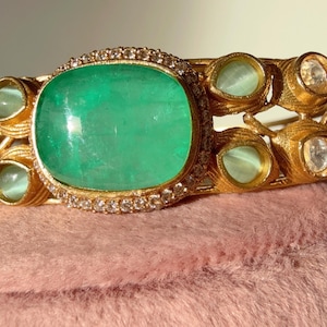 Op de afbeelding: Een goudkleurige armband met een grote, vierkante, smaragdgroene steen in het midden, omringd door kleine diamanten. De armband heeft extra groene en heldere stenen en wordt gepresenteerd op een roze oppervlak. Het ontwerp is sierlijk en gedetailleerd.