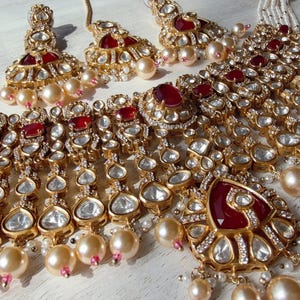 Puede incluir: Un conjunto de joyas dorado con un collar y pendientes a juego. El collar y los pendientes est&aacute;n adornados con piedras preciosas rojas, cristales transparentes y perlas de imitaci&oacute;n. El conjunto tiene un dise&ntilde;o ornamentado.