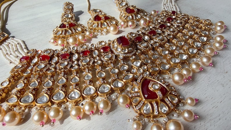 Puede incluir: Un conjunto de joyas dorado con un collar y pendientes a juego. El collar y los pendientes est&aacute;n adornados con piedras preciosas rojas, cristales transparentes y perlas de imitaci&oacute;n. El conjunto tiene un dise&ntilde;o ornamentado.