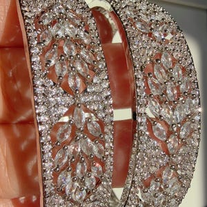 Może przedstawiać: Dwie srebrne bransoletki inkrustowane przezroczystymi, błyszczącymi kryształami. Wzór przedstawia motyw kamieni w kształcie markizy i mniejszych okrągłych kryształów, tworząc olśniewający efekt. Biżuteria jest trzymana na neutralnym tle.