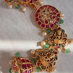 May include: Ornate gold-toned jewelry set with ruby-red and emerald-green accents. The set includes a floral-shaped pendant, a circular pendant, and two smaller pendants with pearl and gemstone embellishments. The jewelry is likely for special occasions.