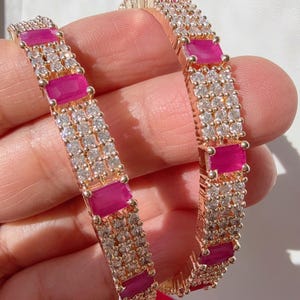 May include: Two rose gold bangles with alternating rows of small clear crystals and rectangular magenta stones. The bangles are held up against a white background.