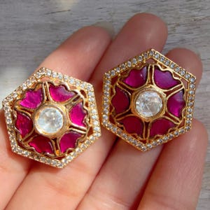 May include: A pair of gold-toned, hexagonal earrings. Each earring features a central, round, clear stone surrounded by a magenta floral design. The outer edge is lined with small, clear stones, creating a sparkling effect. The earrings are held in a hand.