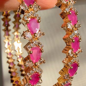 May include: Gold-toned bangles with alternating pink teardrop-shaped gemstones and small white accent stones. The jewelry is set in a textured, ornate design. The bangles are displayed against a neutral background.