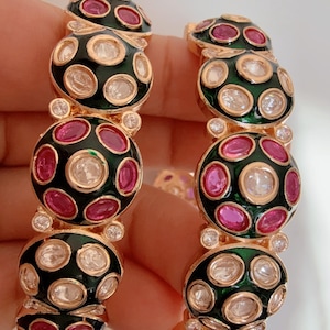 May include: Two gold-toned bangles with a repeating pattern of black enamel circles, each adorned with pink and clear gemstones. The bangles are designed with a series of connected, circular elements, creating a bold and elegant look.