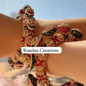 May include: Two gold bangles with red, green, and white enamel and pearl accents. The bangles are stacked on a person's arm. The text "Kundan Creations" is visible on the image.