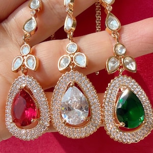 Puede incluir: Tres pendientes colgantes dorados con piedras preciosas en forma de lágrima en rojo, transparente y verde. Cada pendiente presenta un halo de pequeños cristales transparentes alrededor de la piedra preciosa.