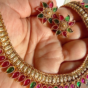 Può includere: Una collana dorata e un set di orecchini abbinati. I gioielli presentano un motivo a goccia ripetuto con pietre preziose rosse e verdi alternate. Gli orecchini sono a forma di fiore, con la stessa combinazione di colori. Il set è esposto su una mano.