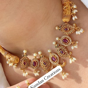 May include: Ornate gold necklace and earrings set with red gemstone centres and small white pearl drops. The jewellery features detailed filigree patterns. The set is displayed on a person, highlighting the craftsmanship and design.