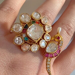 May include: Two gold rings with detailed designs. One ring has a large, clear, round stone surrounded by smaller stones and colourful accents. The other ring is shaped like a peacock, with a clear stone body, ruby accents, and a pearl.