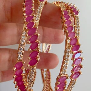 Puede incluir: Dos brazaletes dorados con filas alternas de piedras preciosas rosas con corte marquesa y cristales transparentes con corte redondo. Los brazaletes se muestran sobre un fondo neutro, mostrando el diseño y el color.