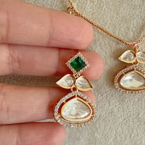 May include: A gold necklace with a green and white gemstone pendant. The pendant features a large, oval-shaped white gemstone surrounded by smaller white gemstones. A smaller green gemstone is set above the oval gemstone.
