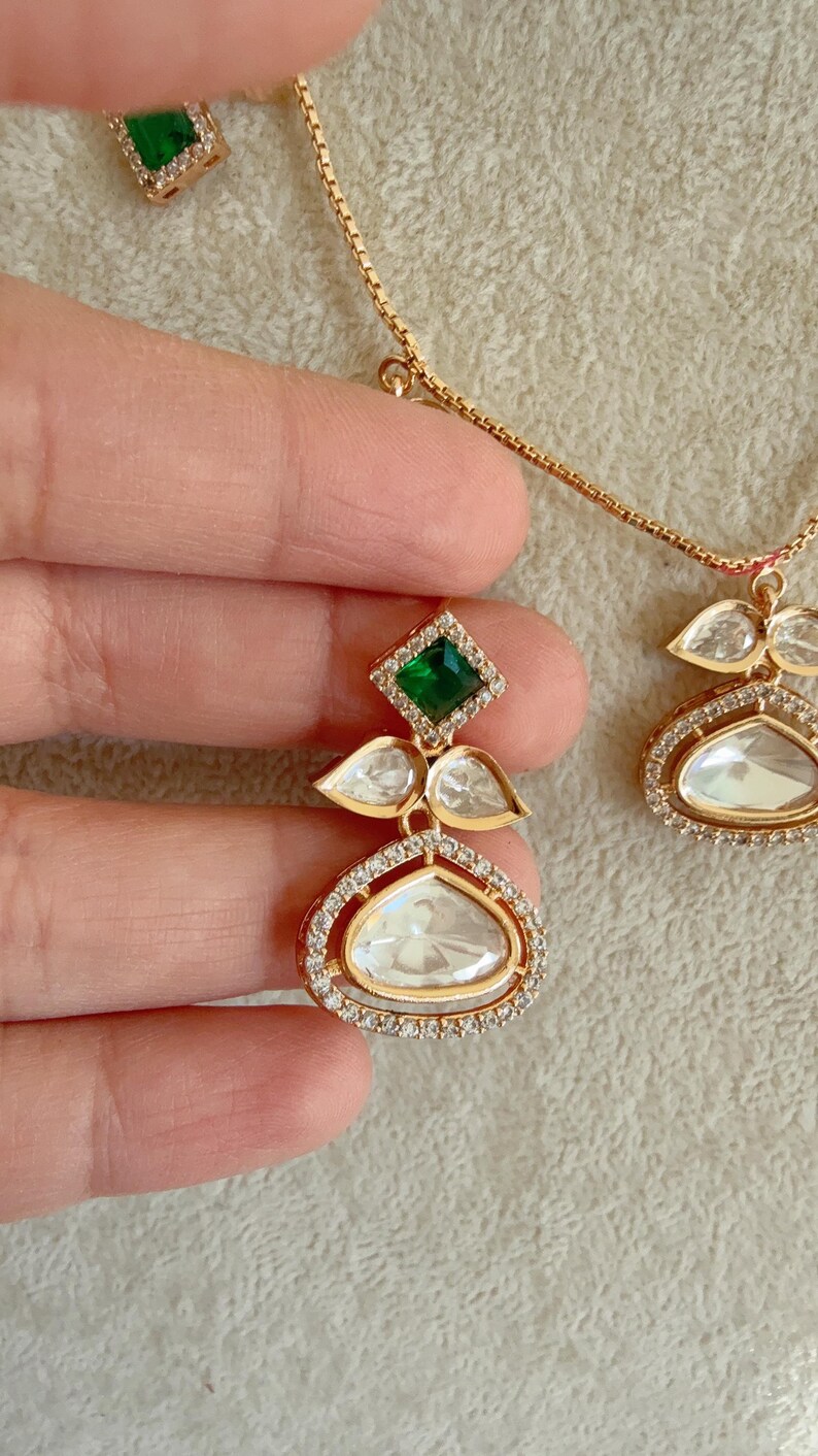 May include: A gold necklace with a green and white gemstone pendant. The pendant features a large, oval-shaped white gemstone surrounded by smaller white gemstones. A smaller green gemstone is set above the oval gemstone.
