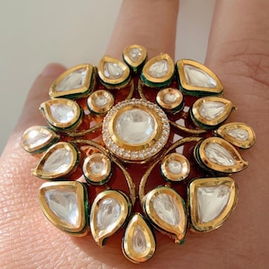 May include: A gold ring featuring a large central clear stone encircled by smaller clear stones and gold detailing. The ring has a floral pattern with teardrop and round stones. The ring is on a finger.