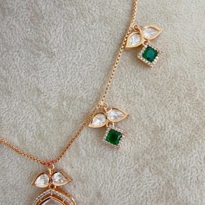 May include: A gold chain necklace with three pendants. Each pendant features a green gemstone surrounded by clear stones and two teardrop-shaped clear stones. The necklace is on a white background.